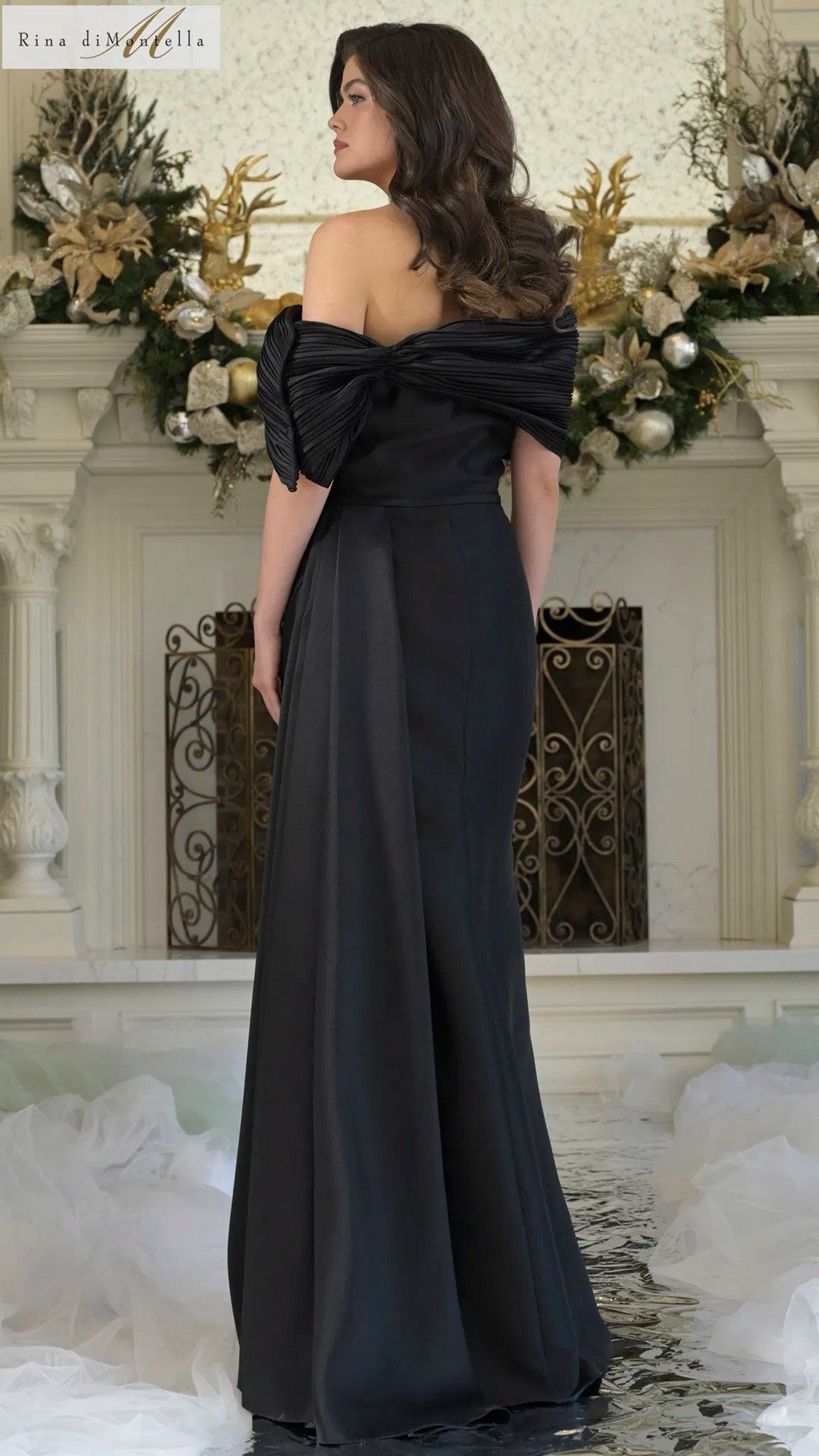 A woman wearing the Rina Di Montella RD3309 Long Formal Dress stands indoors with her back to the camera, the elegant sheath skirt flowing as festive decorations twinkle in the background.